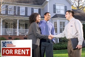 a couple shakes hands with a property manager in front of a house with a "For Rent" sign.